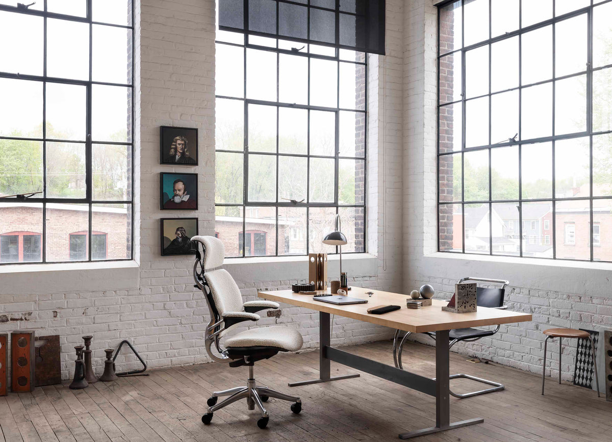 A bright loft-style office setup featuring a white Humanscale Freedom Ocean task chair and an eFloat Go standing desk with a laptop and lamp against a white brick wall background