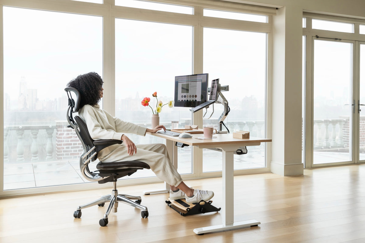 A person working at a Humanscale eFloat Go 2.0 height-adjustable desk on a Freedom 25 Signature Edition chair, featuring a monitor arm with notebook holder, universal accessory bracket, and an FR300 foot rocker in a bright office with large windows