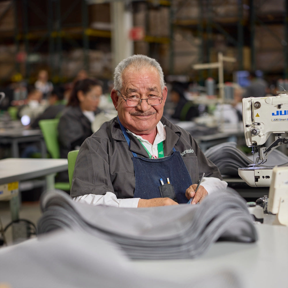A smiling master tailor at a sewing machine with measuring tape and fabric in a busy Humanscale factory