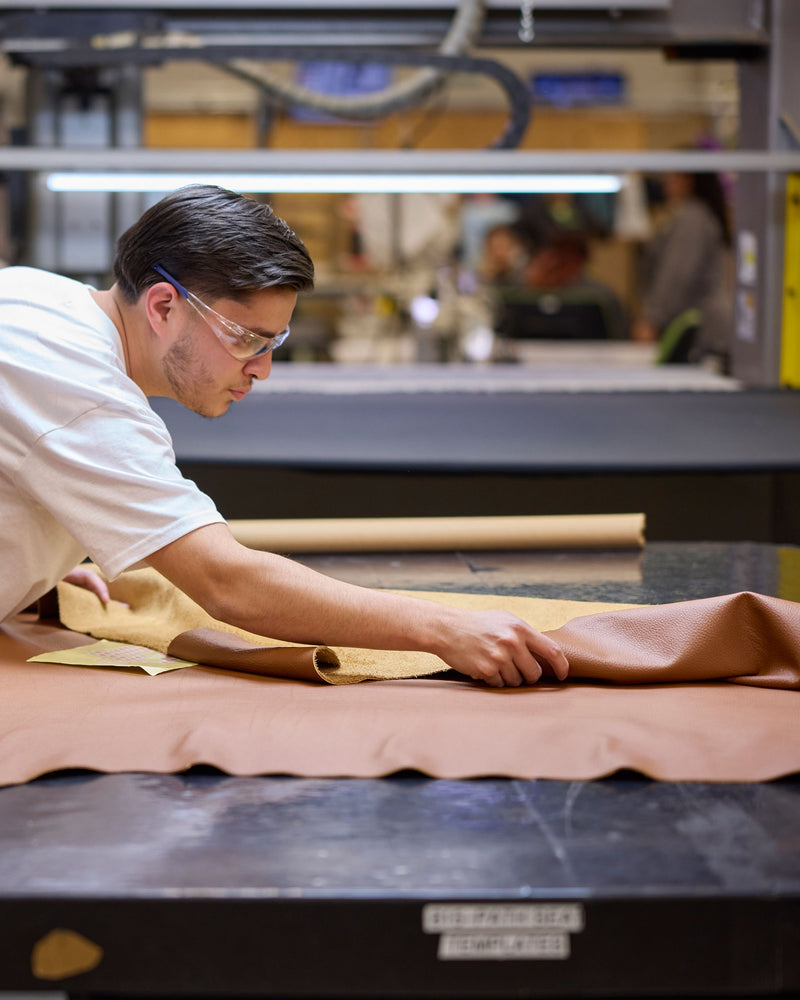 A specialist in a workshop inspecting and smoothing brown leather fabric on an industrial table