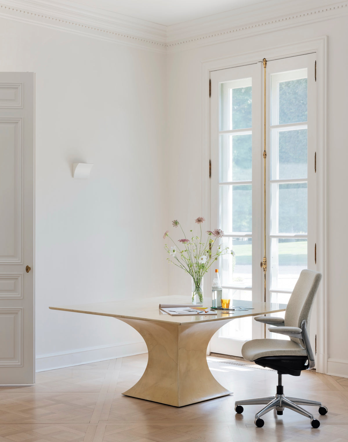 Side profile of a light beige Humanscale Smart Conference chair with a polished aluminum frame next to a wooden desk in a bright room