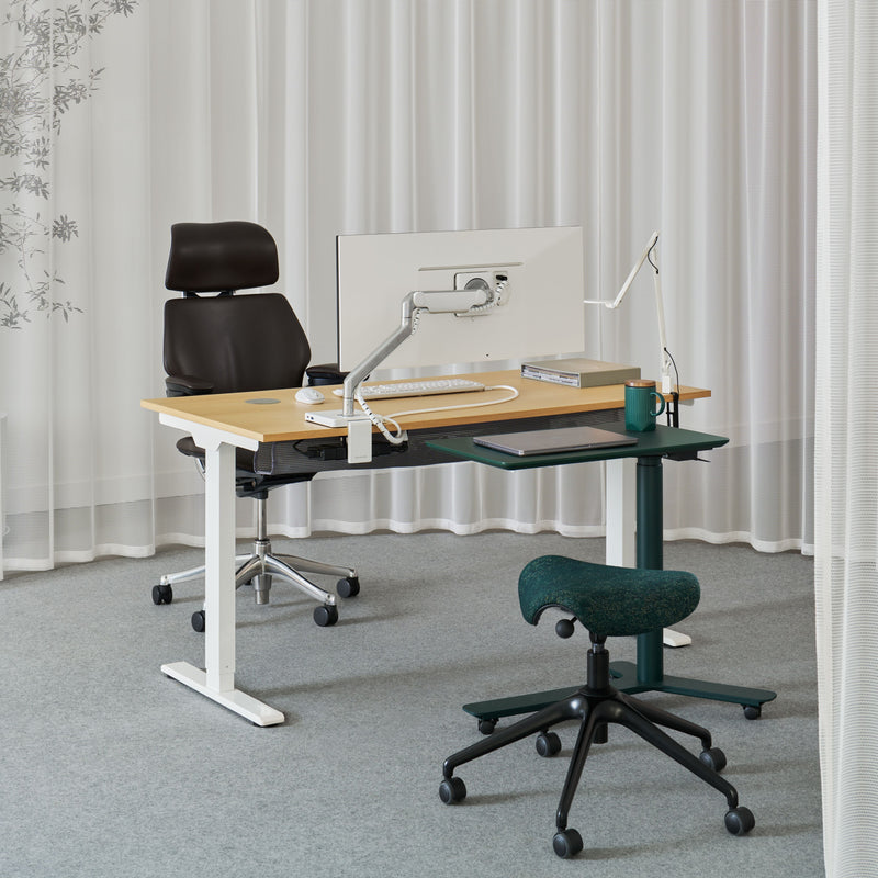 A front view of a modern office setup featuring a Humanscale Freedom Active Pony stool and a Float Mini table in Evergreen, with an eFloat Go 2.0 desk, a white monitor arm, and a Freedom Ocean headrest chair in black saddle leather in the background
