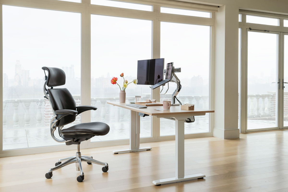 Black Saddle leather Humanscale Freedom Headrest chair next to an eFloat Go 2.0 oak sit-stand desk with a white base, featuring an M8 Pro dual monitor arm and notebook holder with a city view