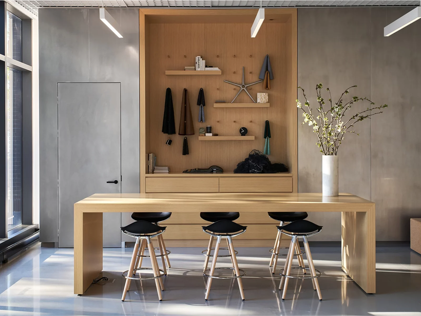 A minimalist modern office featuring six black Humanscale Sedeo bar-height stools with Maple wood legs and polished aluminum frames arranged around a light wood rectangular table