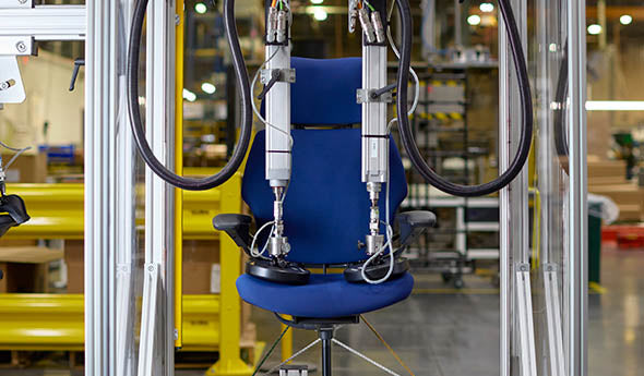 A Humanscale Freedom Ocean Headrest chair undergoing automated durability testing with mechanical arms in a factory setting