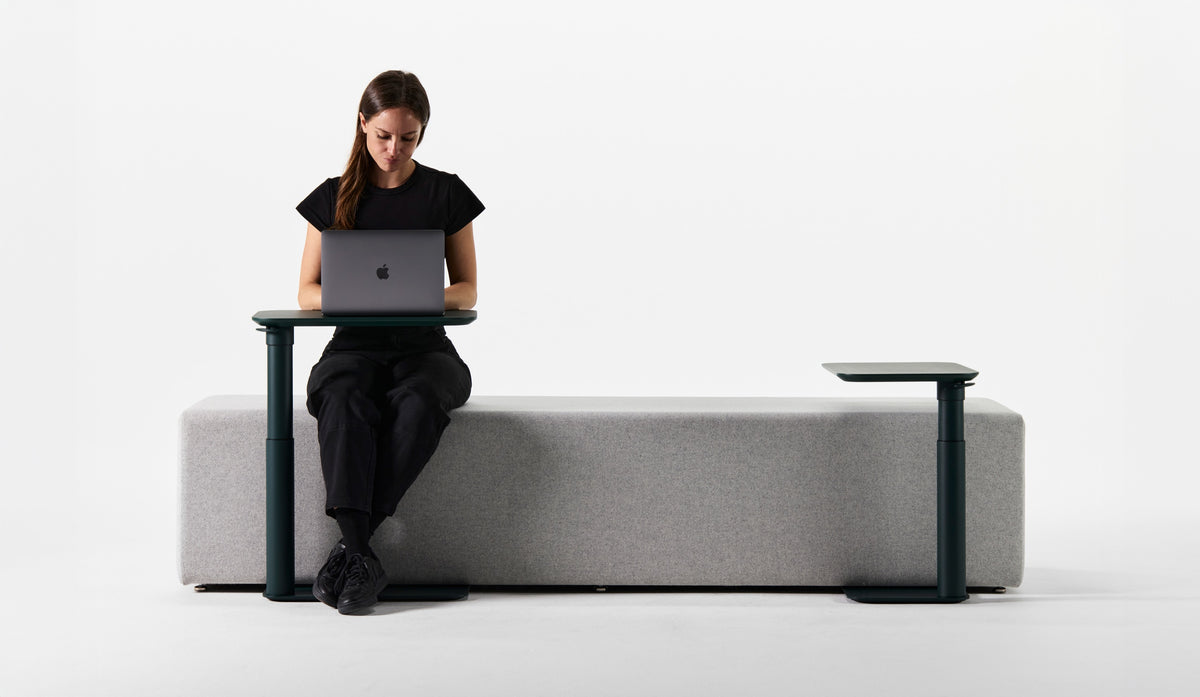 Person using a laptop on a forest green Humanscale Float Micro compact adjustable desk while sitting on a gray bench in a minimalist workspace