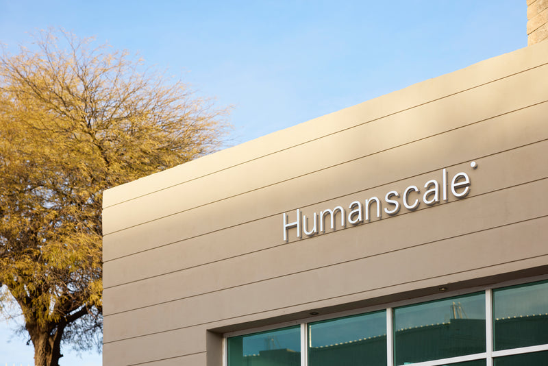 Exterior view of a beige Humanscale building with a silver logo sign and a yellow leaf tree